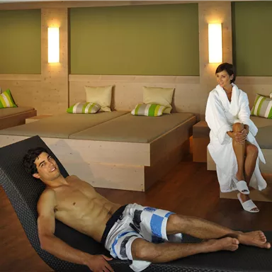 Ein moderner Wellnessbereich mit einem entspannten Mann auf einer Liege und einer Frau in einem Bademantel. Die Umgebung ist hell und freundlich, mit minimalistischer Holz- und Grünausstattung.