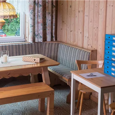 Ein gemütlicher Raum mit Holzwänden und einem Tisch. Im Hintergrund gibt es eine Fensterbank mit einer Lampe und eine Sitzbank.