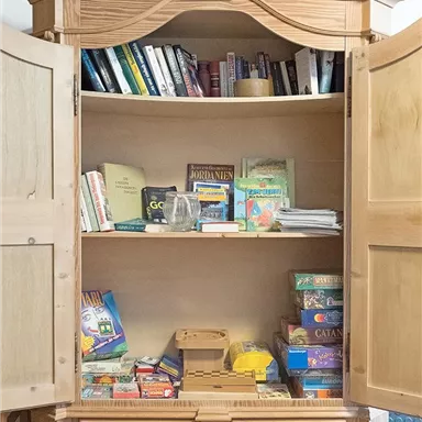 Ein schöner Holzschrank mit offenen Türen, der Bücher und Gesellschaftsspiele enthält. Auf den Regalen sind verschiedene Spiele und Lehrbücher ordentlich sortiert.