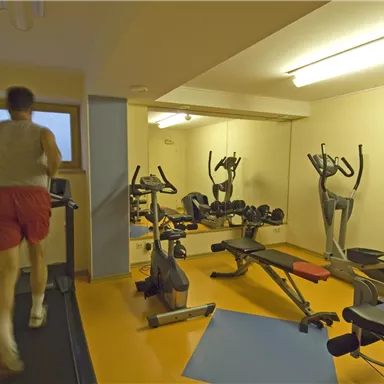 Ein Fitnessraum mit einem Laufband und verschiedenen Trainingsgeräten. Eine Person joggt auf dem Laufband.
