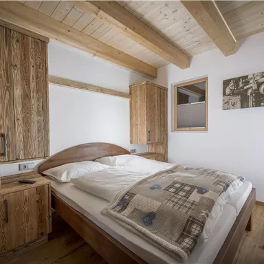 A cozy bedroom with a large bed and wooden shelves. The walls are painted light and there is a window that lets in natural light.
