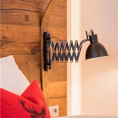 Eine elegante Wandlampe ist an einem Holzpaneel befestigt, das mit einem Schieferrand dekoriert ist. Im Vordergrund liegt ein rohes Kissen mit einem dekorativen Design.