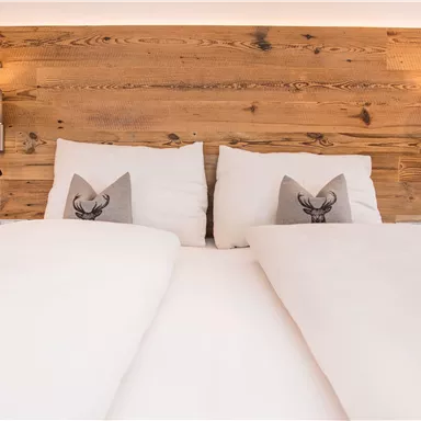 Ein gemütliches Doppelbett mit weißen Bettdecken und dekorativen Kissen. Im Hintergrund befindet sich eine Holzverkleidung mit warmen Lichtquellen.