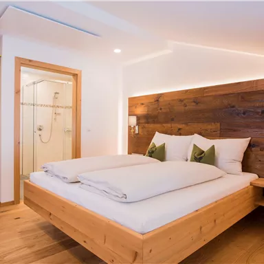 Ein modernes Schlafzimmer mit einem Doppelbett aus Holz und stilvollen Kissen. Im Hintergrund ist ein Bad mit Glasduschkabine zu sehen.