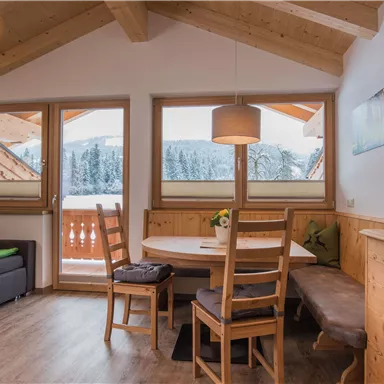 Ein gemütlicher Wohnraum mit Holzmöbeln und großen Fenstern. Draußen ist ein schneebedeckter Ausblick auf die winterliche Landschaft.