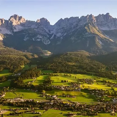 Eine beeindruckende Berglandschaft mit hohen Gipfeln und grünen Wiesen. Im Vordergrund sind kleine Dörfer und eine malerische Natur zu sehen.