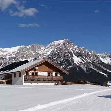 Eine gemütliche Berghütte steht in einer verschneiten Landschaft. Im Hintergrund erheben sich beeindruckende Berge unter einem klaren blauen Himmel.