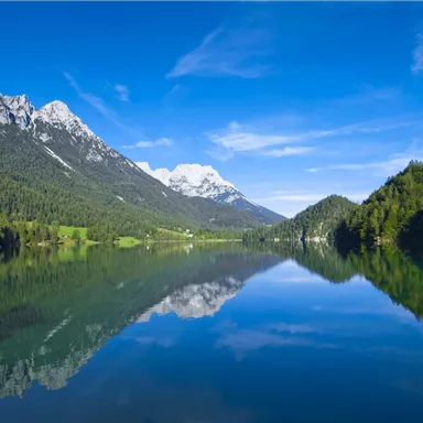 Ein schöner See umgeben von Bergen und Bäumen. Der klare Himmel spiegelt sich im Wasser.