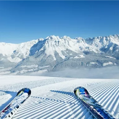 Eine winterliche Berglandschaft mit schneebedeckten Gipfeln und klarblauem Himmel. Im Vordergrund liegen Skier auf frisch präparierter Piste.