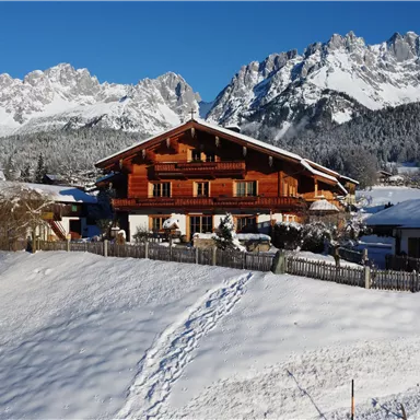 A charming wooden house sits on a snow-covered hill. In the background, majestic mountains stretch under a clear blue sky.