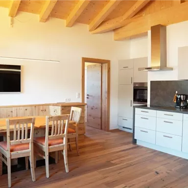 Eine moderne Küche mit Holzboden und offenen Regalen. Der Essbereich hat einen großen Tisch und Stühle, und an der Wand hängt ein TV.
