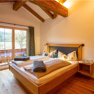 Ein gemütliches Schlafzimmer mit einem Doppelbett und Holzmöbeln. Große Fenster bieten Blick auf den Balkon und die Natur.