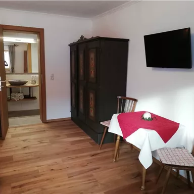 A cozy room with wooden floors and bright walls. There is a table with a red table runner, chairs, and a television on the wall.