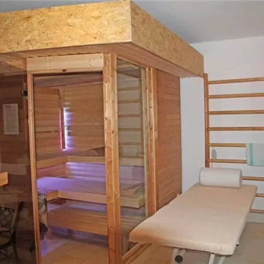 A stylish sauna room with a glass front and wooden details. Next to the sauna, there is a lounger and a rack for towels.