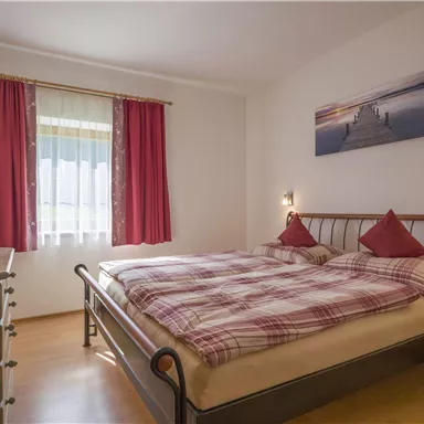Ein gemütliches Schlafzimmer mit einem großen Bett und beige-roter Bettwäsche. Die Fenster haben helle Vorhänge und der Raum ist mit Holz- und Dekorationsmöbeln ausgestattet.