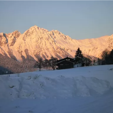 Eine verschneite Landschaft mit schneebedeckten Bergen im Hintergrund. Im Vordergrund steht ein Holzhaus, umgeben von Schnee.