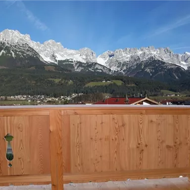 Eine Holzbalustrade mit Blick auf hohe Berge, die mit Schnee bedeckt sind. Der Himmel ist klar und blau, und im Vordergrund sind einige alpine Häuser sichtbar.