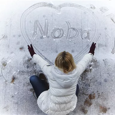 A person is sitting on a frozen ground and has drawn a heart with the name "Noba" in the snow. They are wearing a white jacket and red gloves.