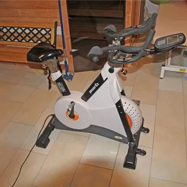 A stationary bike is located in a room with tiled flooring. In the background, a bench and a table can be seen.