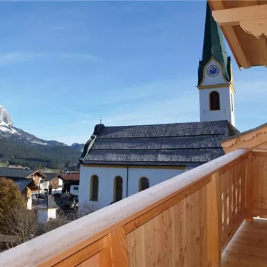 Ein Blick auf eine Kirche mit einem hohen Turm und schneebedeckten Bergen im Hintergrund. Auf der Vorderseite ist ein Holzgeländer zu sehen.