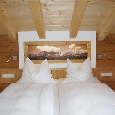 Ein gemütliches Schlafzimmer mit Holzdecke und einem Doppelbett. Im Hintergrund befindet sich ein wunderschönes Bergbild.