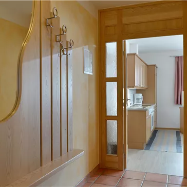 A bright hallway with wooden panels and a coat rack. In the background, an open kitchen is visible.