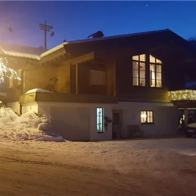 A cozy wooden house in the snow, illuminated by lights in the twilight. The surroundings are wintry and peaceful.