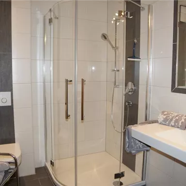 Ein modernes Badezimmer mit einer Duschkabine und einem Waschbecken. Die Wände sind hell gefliest und es gibt einen großen Spiegel.