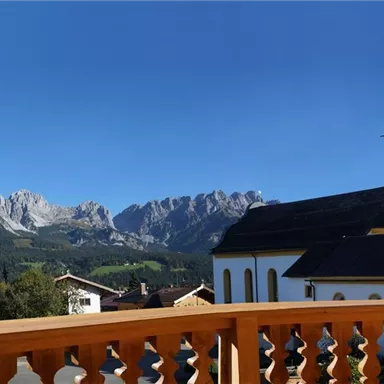 A picturesque landscape with a church tower and majestic mountains in the background. The sky is clear and blue.