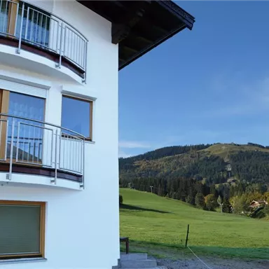Ein modernes Gebäude mit einem großen Balkon und Fenstern. Im Hintergrund erstreckt sich eine grüne Wiese und sanfte Hügel unter einem klaren blauen Himmel.