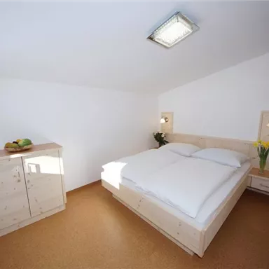 Ein modernes Schlafzimmer mit einem Doppelbett und einer hellen, minimalistischen Einrichtung. Auf dem Nachttisch stehen frische Blumen und ein Obstkorb.