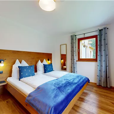 A cozy bedroom with two beds and blue pillows. There are large windows that provide plenty of daylight.