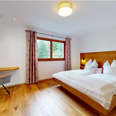 A modern bedroom with a large bed and wooden details. The room has large windows and is decorated in a friendly and bright manner.