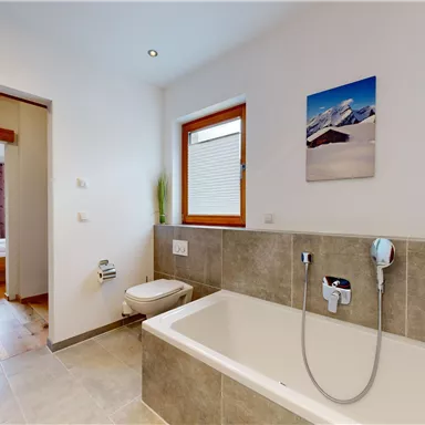 A modern bathroom with a bathtub and a shower. In the background, a cozy bedroom is visible.