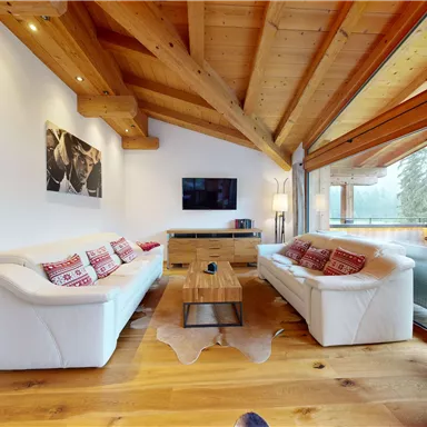 A cozy living room with modern white sofas and a wooden table. Large windows and a wooden ceiling create a bright and inviting atmosphere.