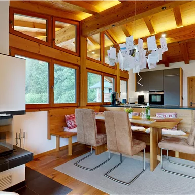 A modern, bright living room with a wooden ceiling and large windows. The dining area is stylishly furnished with a table and beige chairs.