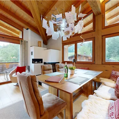 A cozy living space with a wooden beam ceiling, large windows, and a view into the greenery. The table is decorated with drinks and a red blanket.
