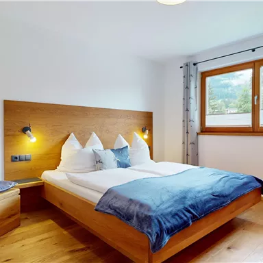 A bright bedroom with a double bed and modern wooden furniture. The room has large windows that let in daylight.