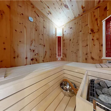 A bright wooden sauna with benches and a heating stove. The walls are clad in wood, and there are small windows for light.