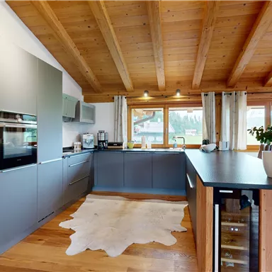 A modern kitchen with a wooden ceiling and bright floor. The kitchen unit is gray and offers plenty of storage space as well as windows with a view outside.