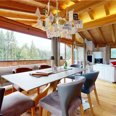 A modern and bright living room with a wooden ceiling and large windows that offer a view of nature. The dining table is elegantly set, surrounded by stylish chairs.