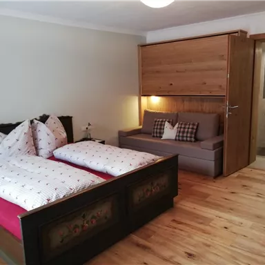 A cozy bedroom with two beds and a sofa. The floor is made of wood, and there is warm light as well as wooden details in the room.