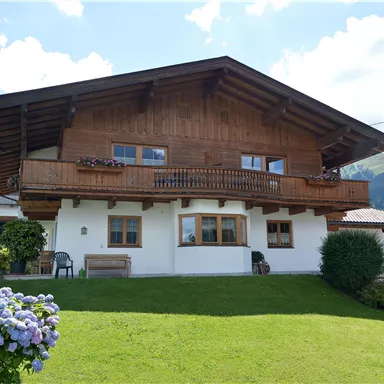 A traditional alpine wooden house with a balcony. The garden is well-maintained and colorful flowers adorn the foreground.