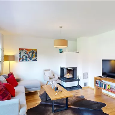 A modern living room with a cozy sofa and a wood stove. The room is bright and friendly, with a large TV and a small table.