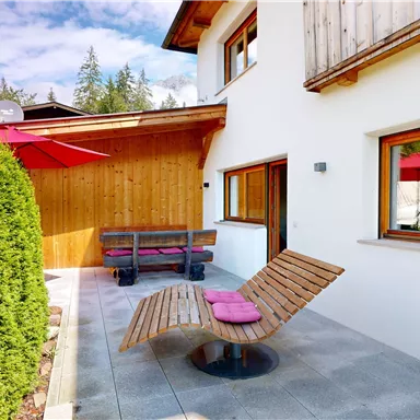 A cozy outdoor area with modern furniture and a privacy screen made of hedges. Umbrellas provide shade and an inviting atmosphere.