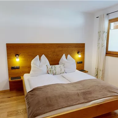 A cozy bedroom with a wooden bed and two pillows. The room has large windows and a modern, bright decor.