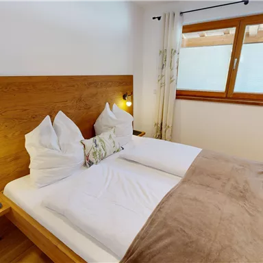 A cozy bedroom with a double bed and wooden details. Large windows provide plenty of daylight.