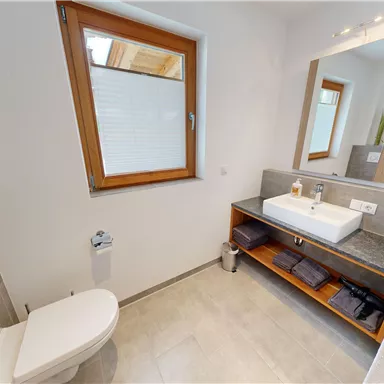 A modern bathroom with a sink, mirror, and a shower. The windows provide plenty of natural light and there are neatly folded towels.