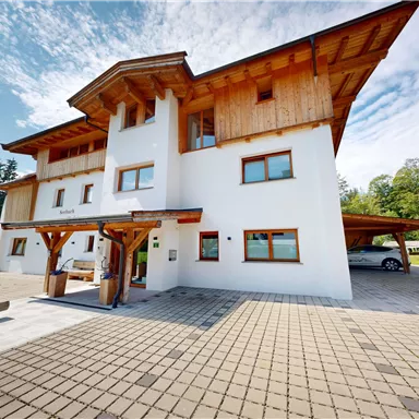 A modern building with wooden details and large windows. Surrounded by a green meadow and trees under a clear sky.