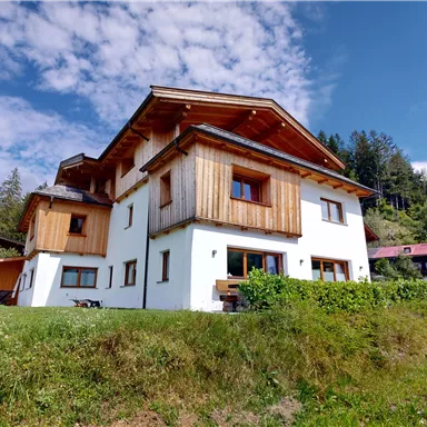A modern house with wooden cladding and a beautiful garden. In the background, green hills and a clear sky can be seen.